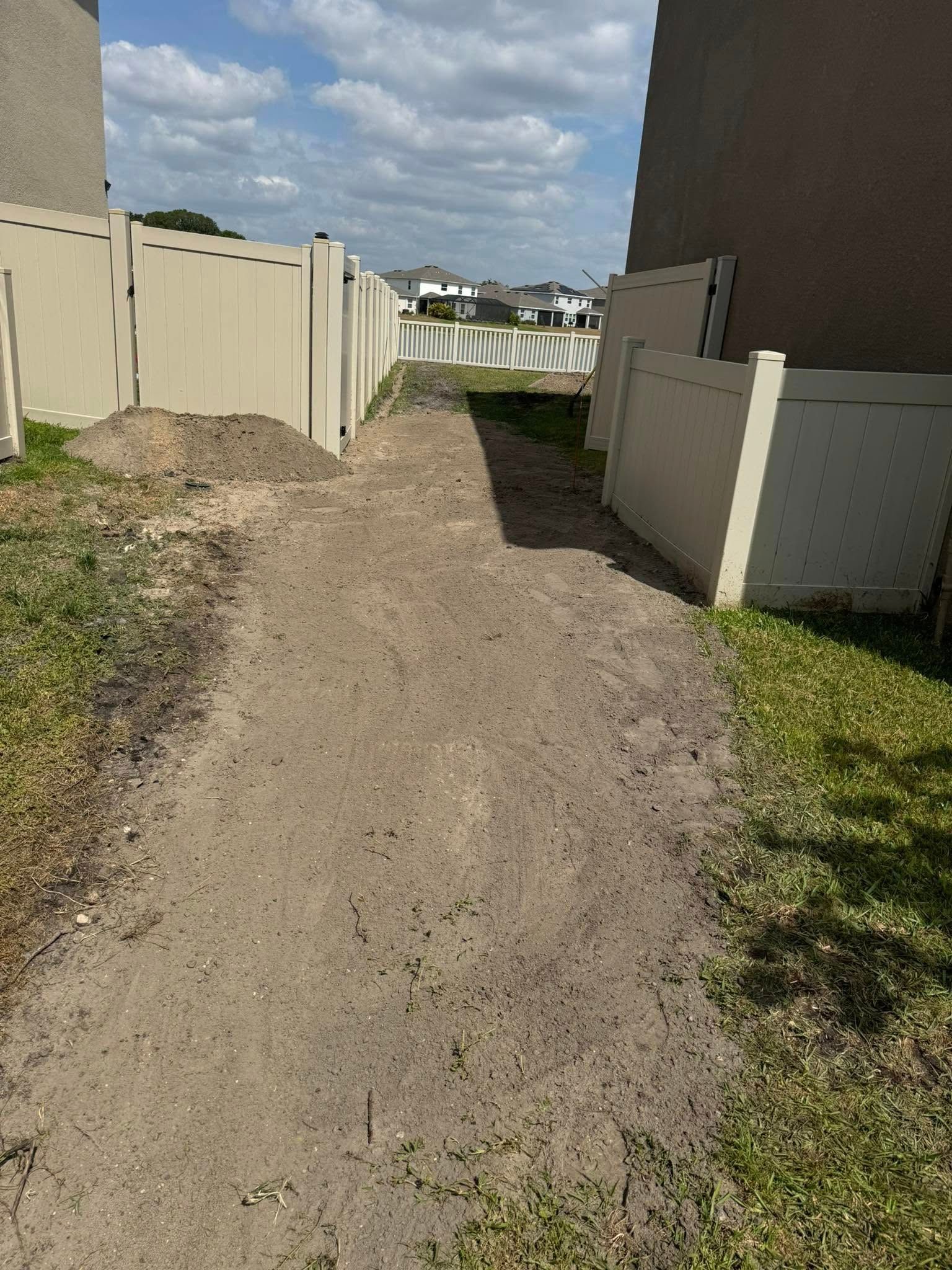 A narrow dirt path between two beige fences, leading towards a distant neighborhood on a sunny day.