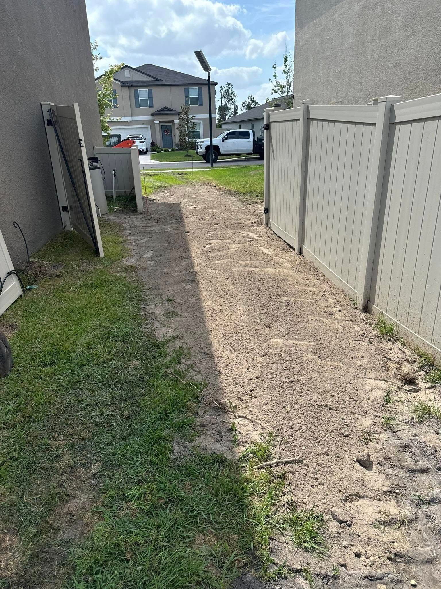 A narrow dirt path between two fences and a building leads to a street with houses.