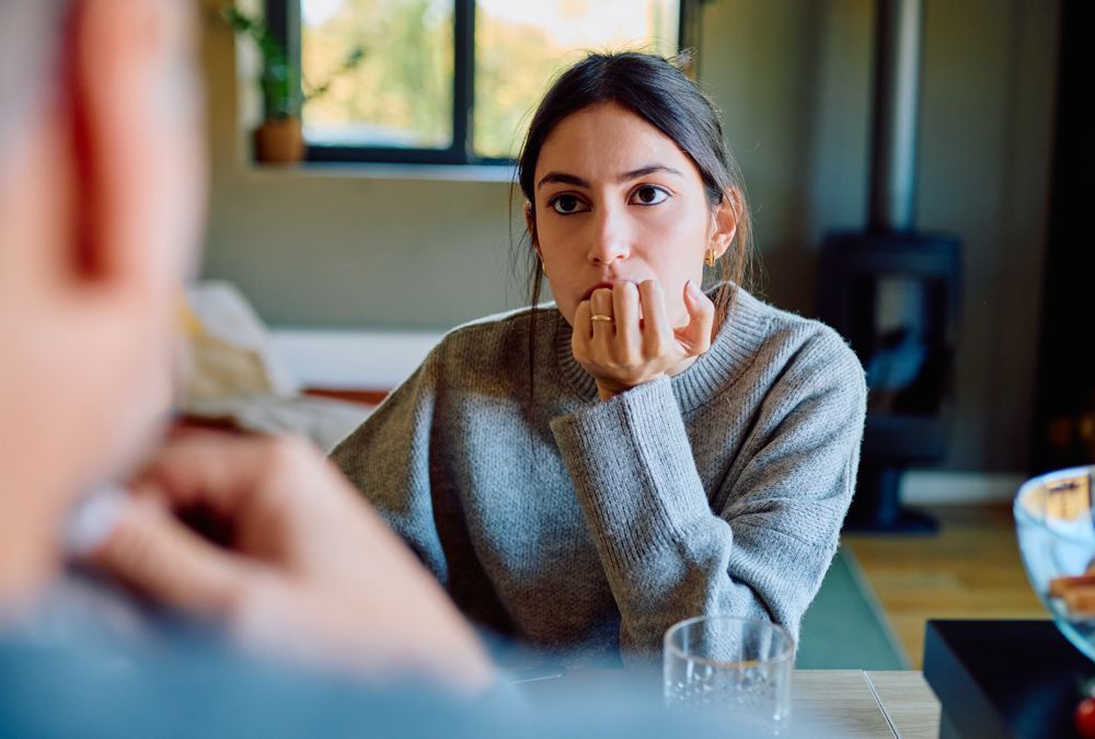 Woman in gray sweater listening thoughtfully at a table in a bright café.
