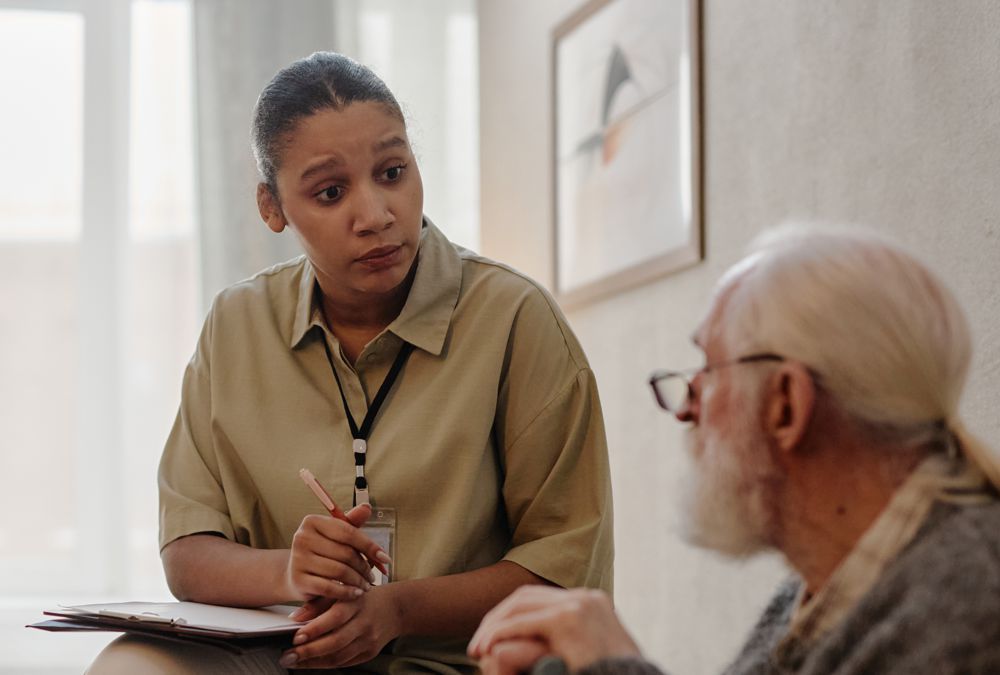 A caregiver speaks with an elderly man at a table, holding a pen and notes in a bright room.