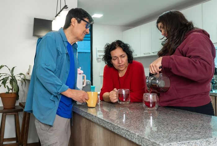 Three people in a kitchen preparing drinks, with one pouring coffee into a glass on the counter.