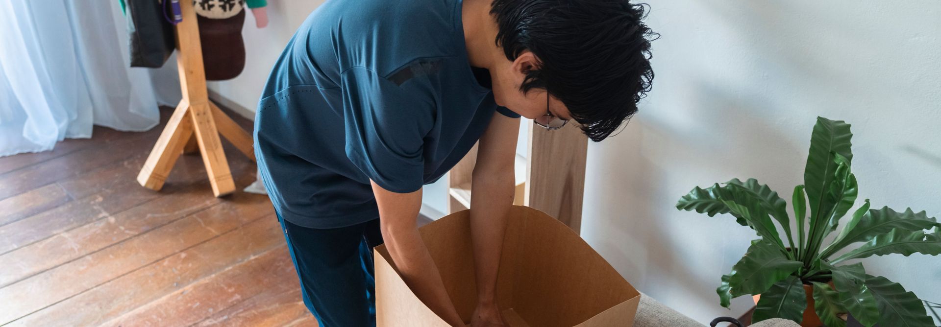 Person bending over an open cardboard box in a room with wooden floor and potted plant nearby
