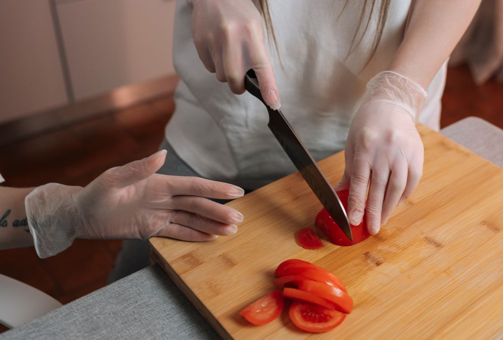 Hands in gloves slicing red tomato on a cutting board with a knife