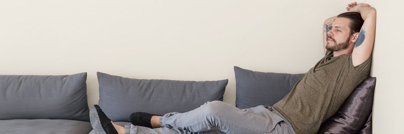 Man reclining on a gray couch, looking relaxed with hands behind his head.