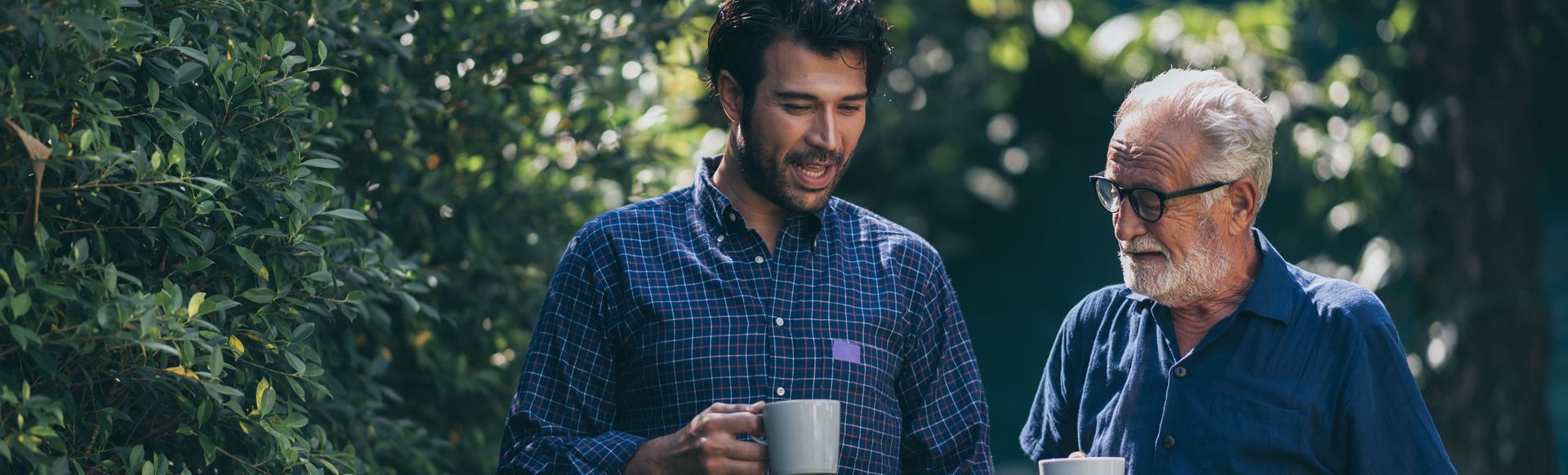 Two men standing outdoors, smiling and holding mugs in a leafy garden