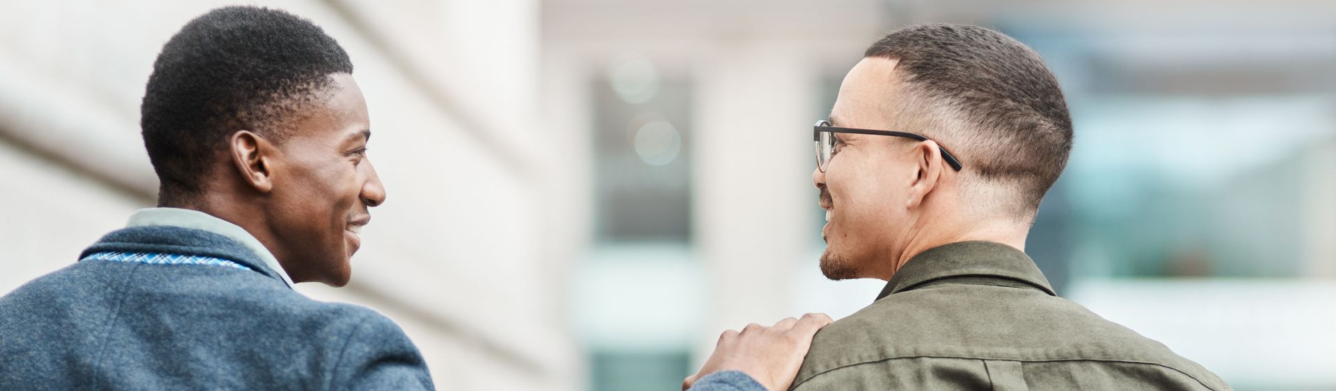 Two people smiling and talking outdoors, seen from behind in casual jackets