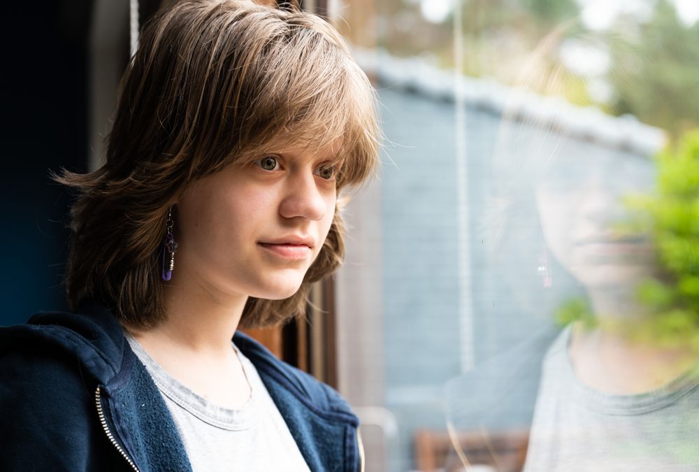 Person with shaggy brown hair looking out a window, wearing a dark hoodie and white shirt.