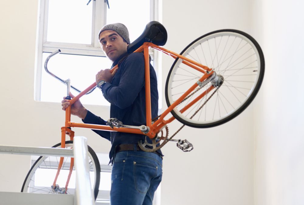 Person carrying an orange bicycle up a staircase indoors, near a bright window.