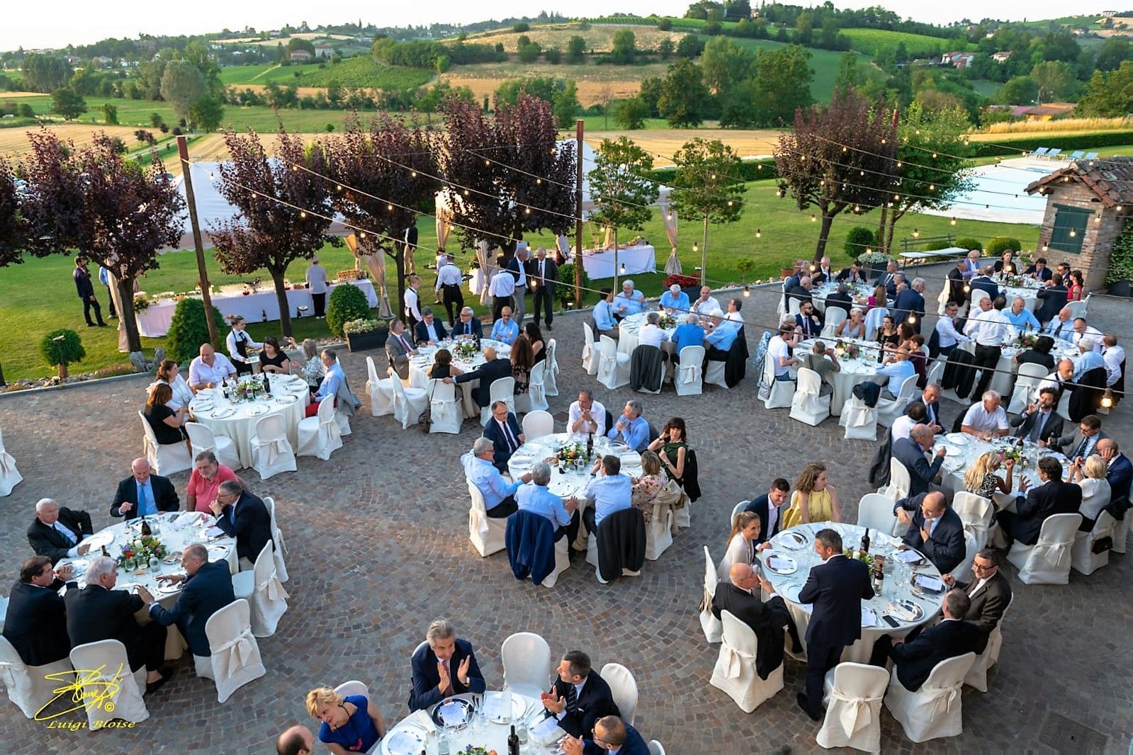 Banchetto all'aperto con gli ospiti seduti a tavoli rotondi con vista su un pittoresco paesaggio rurale.