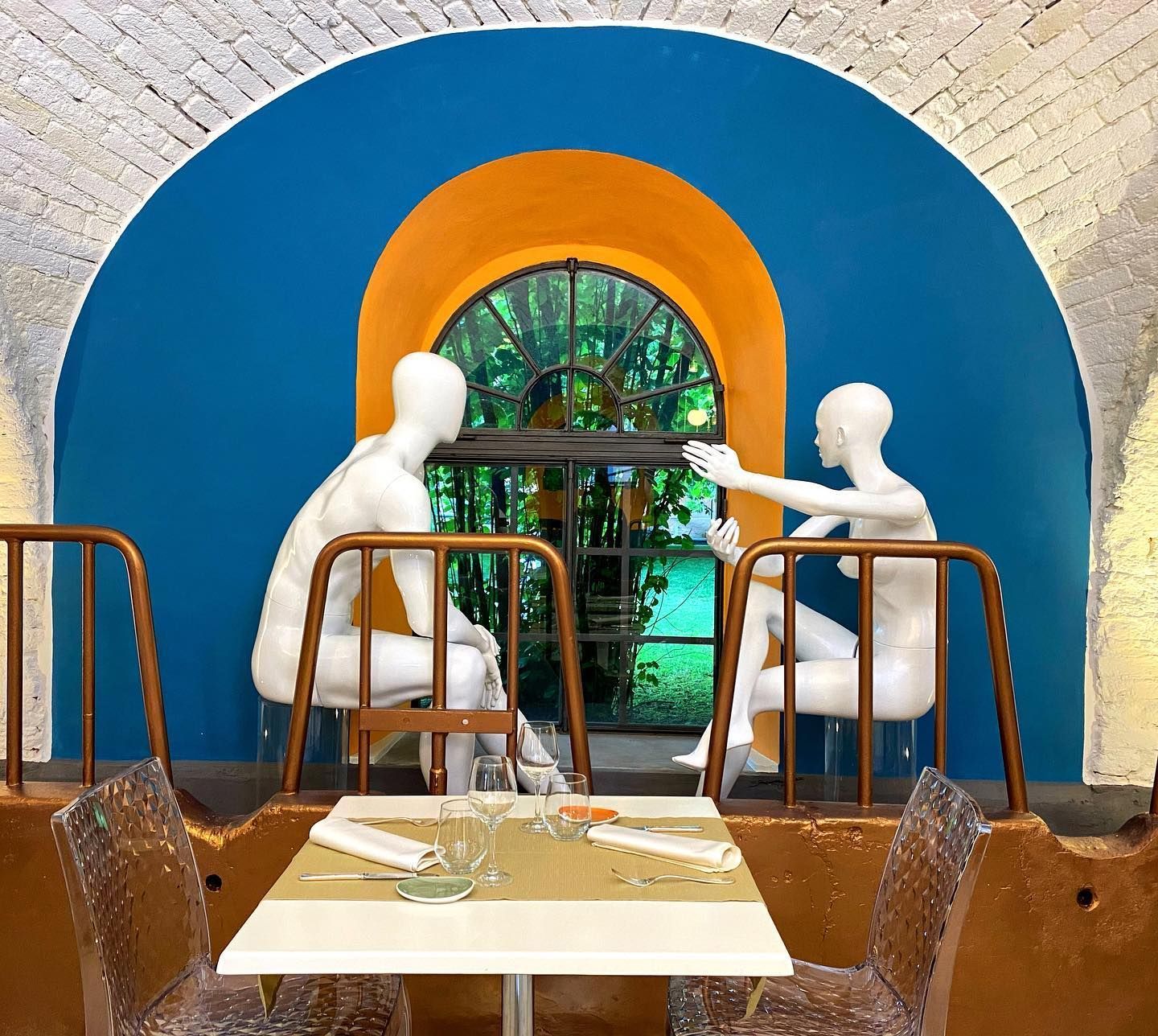 Due manichini bianchi sono seduti a un tavolino da caffè in una nicchia ad arco blu e arancione.