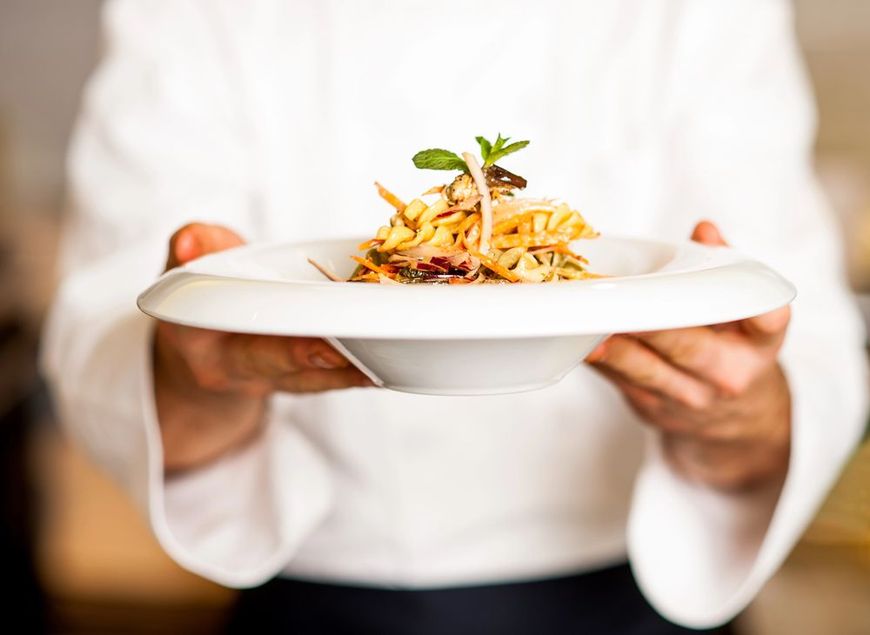 Persona che tiene in mano un piatto bianco di pasta guarnita con erbe aromatiche e verdure.