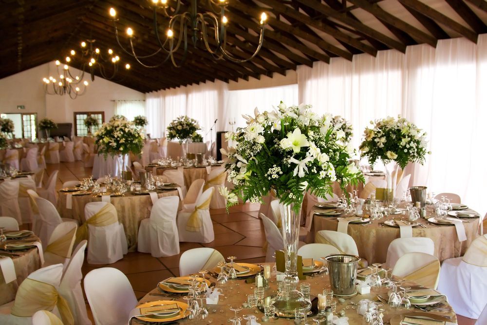 Elegante sala ricevimenti di nozze con tavoli rotondi, coprisedie bianchi e alti centrotavola floreali.