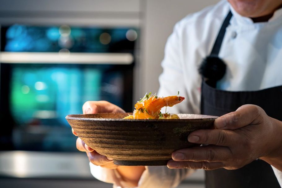 Uno chef tiene in mano un piatto gourmet in cucina, con un forno sfocato sullo sfondo.