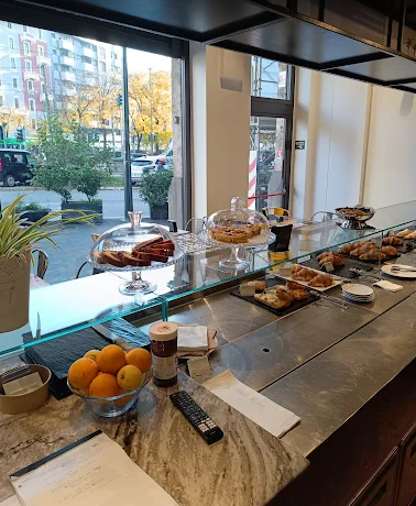 Colazione a buffet in hotel con pasticcini, frutta e bevande su un bancone vicino alle grandi finestre.