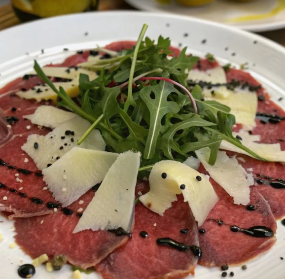 Carpaccio di manzo tagliato a fettine sottili, guarnito con rucola, formaggio grattugiato, semi di sesamo nero e un filo di aceto balsamico, servito su un piatto bianco.