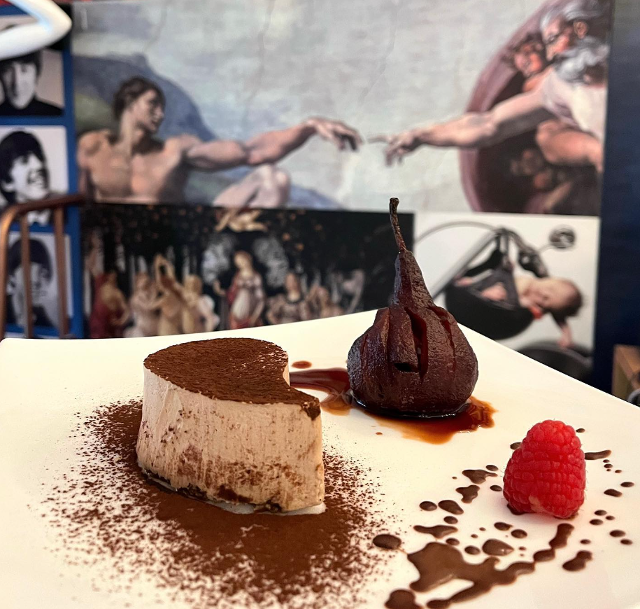 Piatto da dessert al cioccolato con tiramisù, pera sciroppata, lampone e salsa su un piatto bianco.