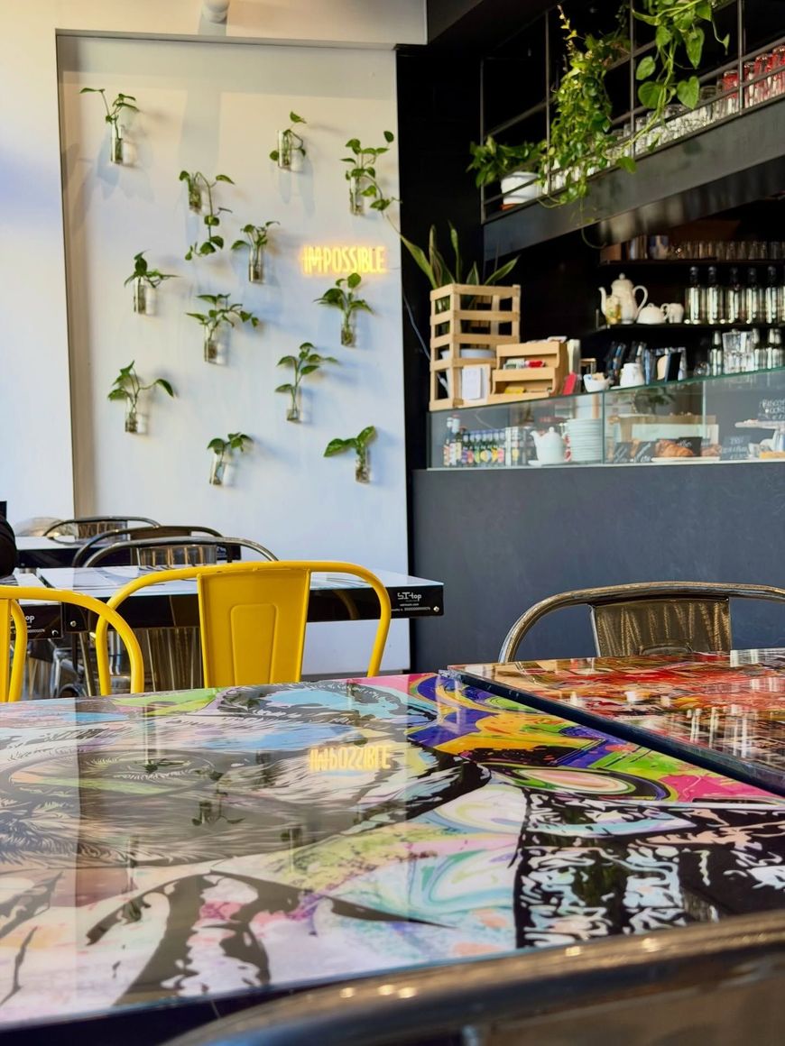 Interno colorato di un caffè con piante alle pareti, sedie gialle e un tavolo dipinto lucido in primo piano.