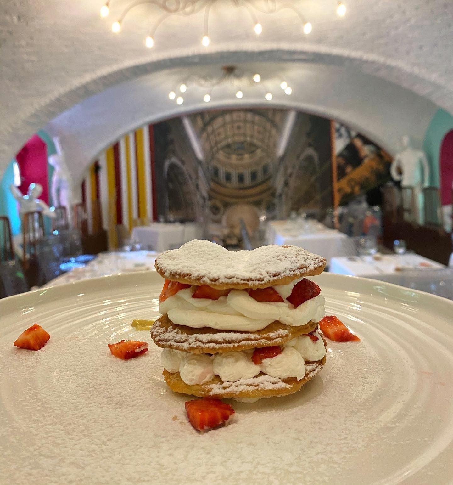 Torta di pasta frolla con zucchero a velo, panna e fragole, servita su un piatto in un caffè illuminato da luci soffuse.