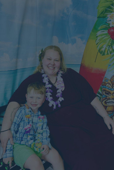 Woman and child sitting together, smiling, with a blue ocean backdrop and flower leis.