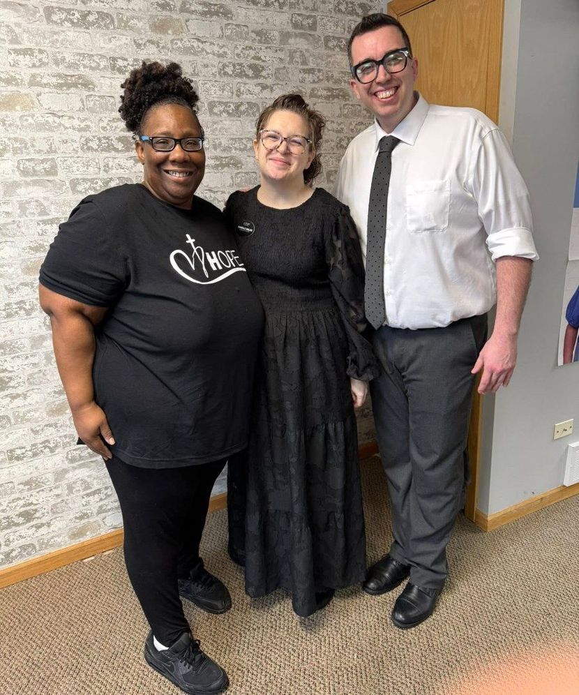 Three people standing together indoors, smiling, with a wall and carpeted floor behind them.