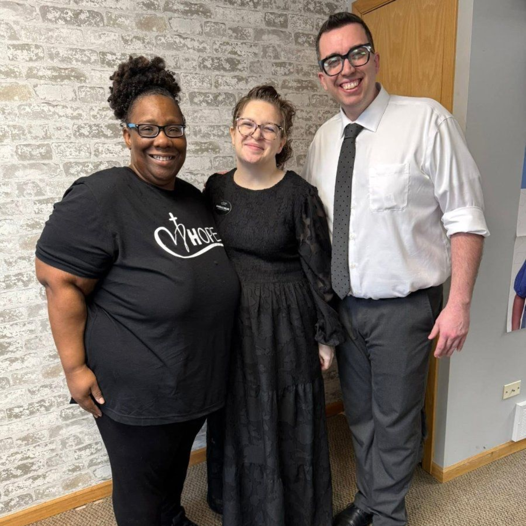 Three people smiling indoors, standing together against a brick wall.
