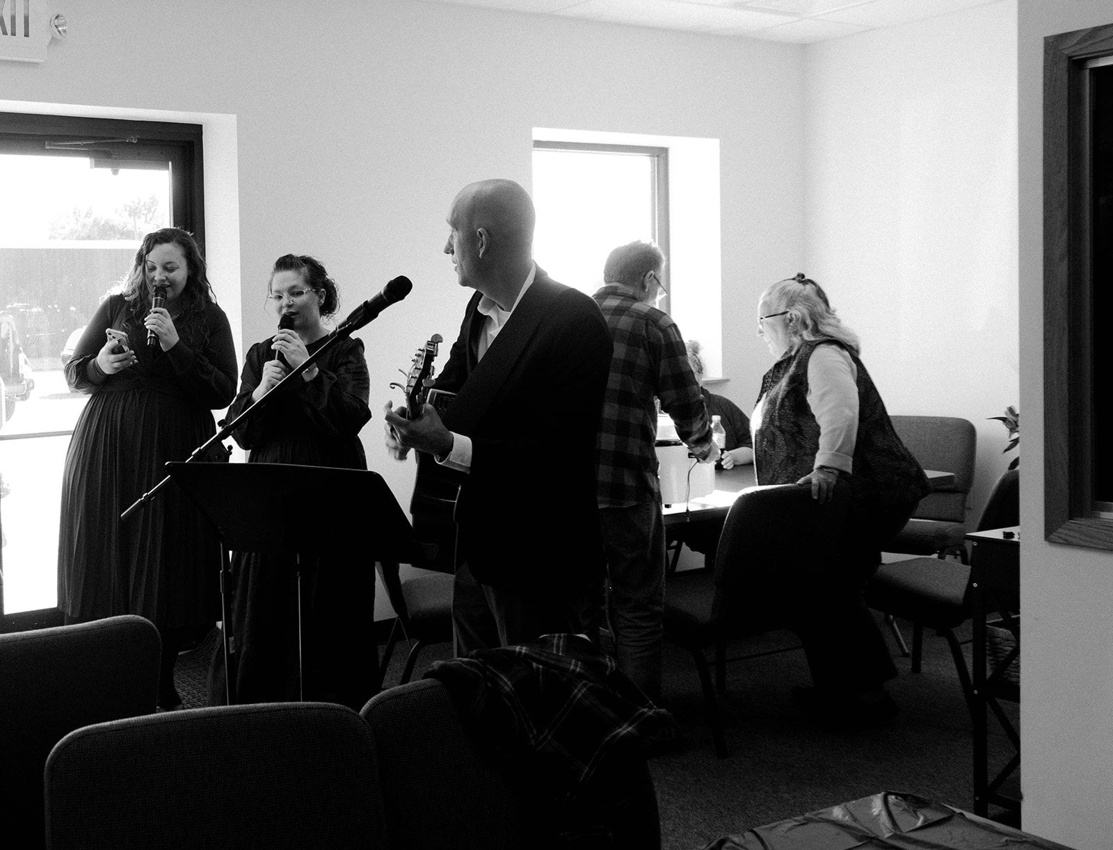 Musicians performing in a bright room, with guitar, violin, and seated players near a window.