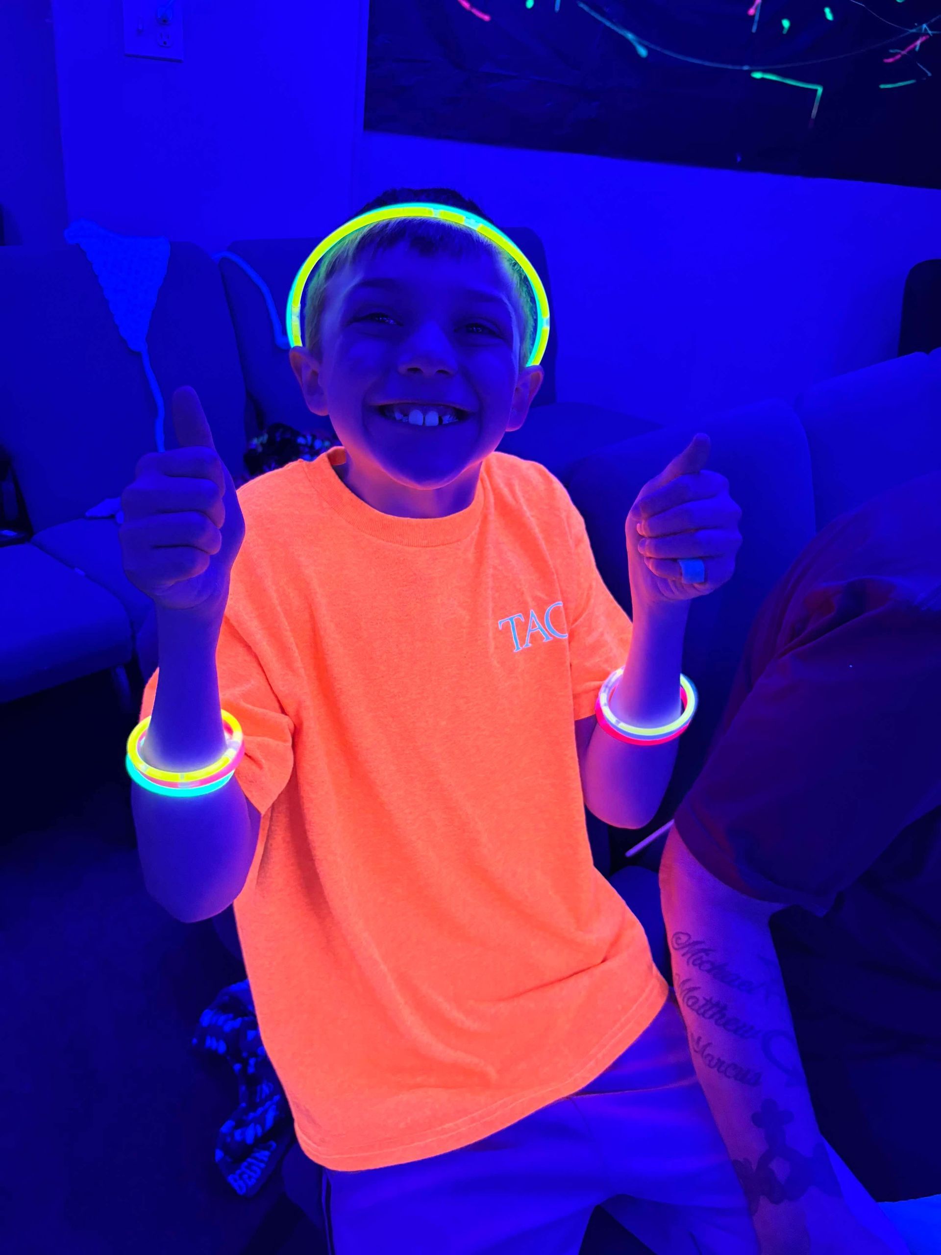 Child in neon orange shirt smiling and giving two thumbs up under blue-black light, wearing glowing bracelets.