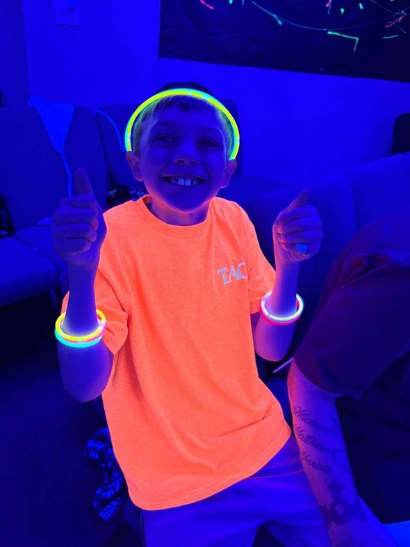 Child in a neon orange shirt smiling and giving two thumbs up under blue lighting, wearing glow bracelets.