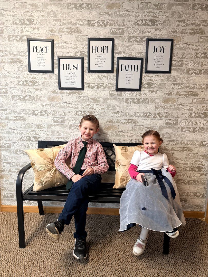 Two children sitting on a bench in front of a patterned wall with framed art above them.