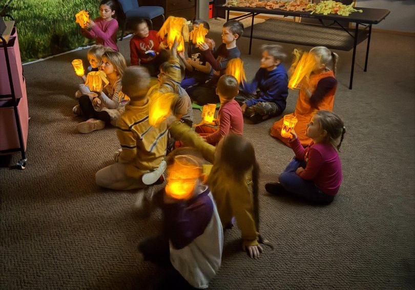 Children seated in a circle on a carpet, holding glowing lanterns in a classroom.