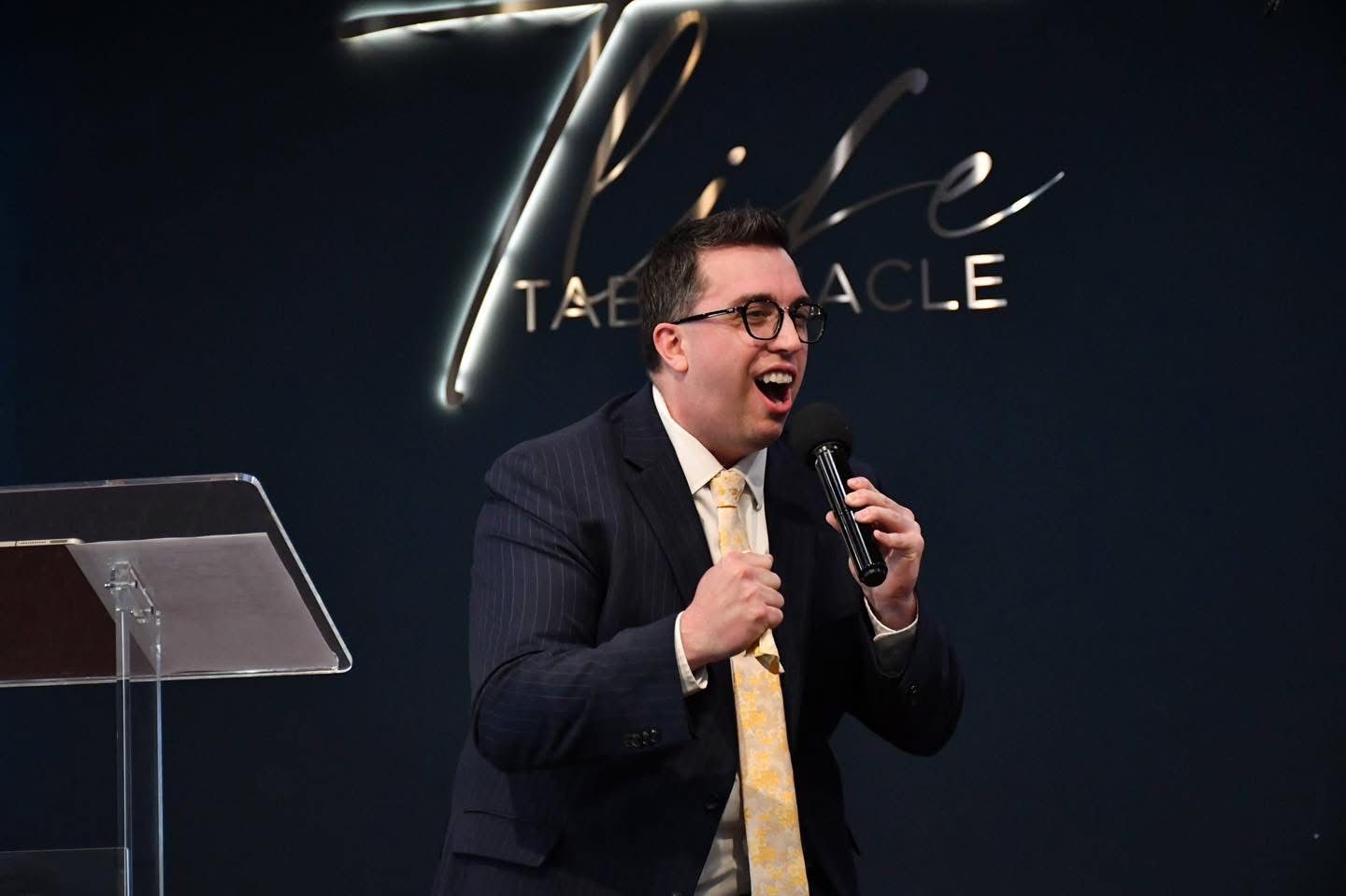 Man speaking passionately into a microphone on a stage with a podium and sign behind him