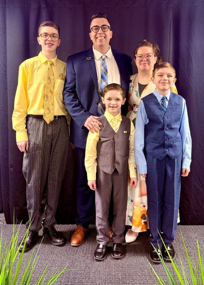Five people posing formally indoors; one man in a navy suit stands centered behind four children in bright outfits.