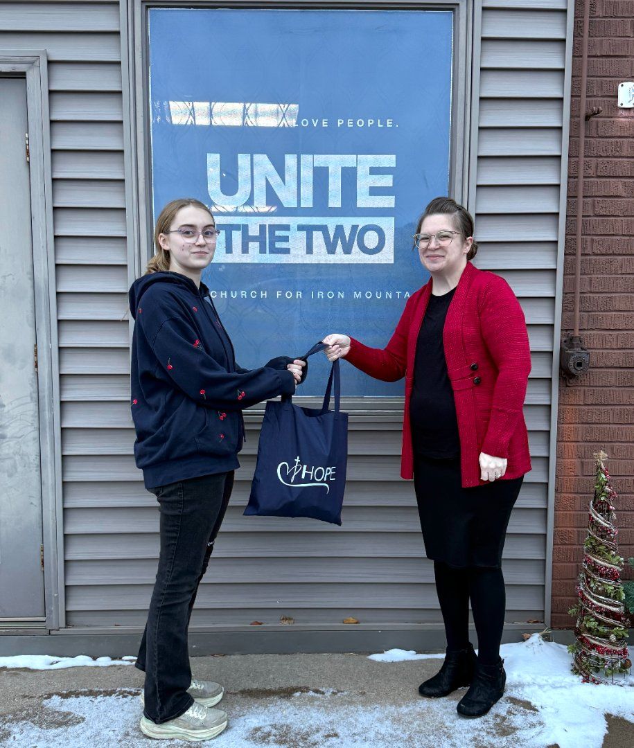 Two people exchanging a gift bag in front of a blue UNITE THE TWO sign