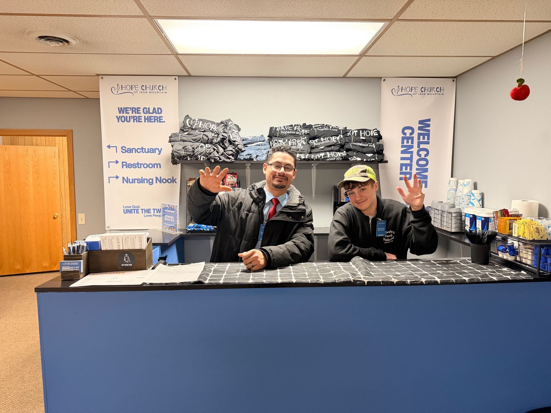 Two people behind a counter in a coffee shop, smiling and waving under a welcome sign.
