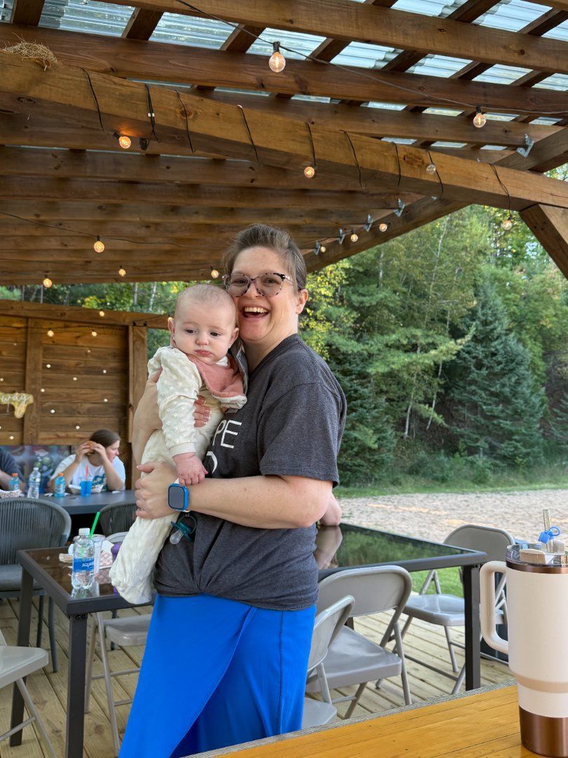 Person holding a baby under a wooden pavilion with picnic tables and trees in the background