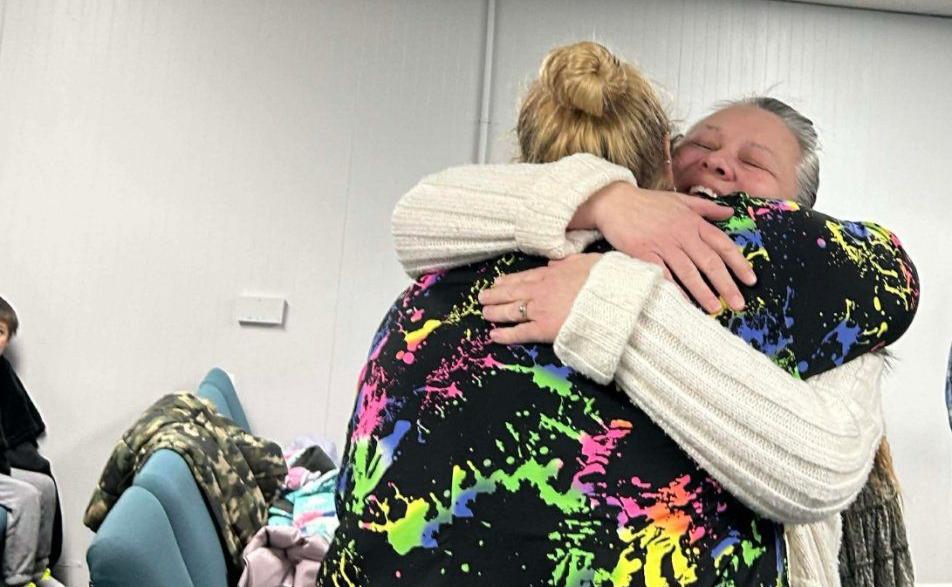 Two people hugging in a bright room, one in a black neon-patterned sweater and the other in a white sweater.