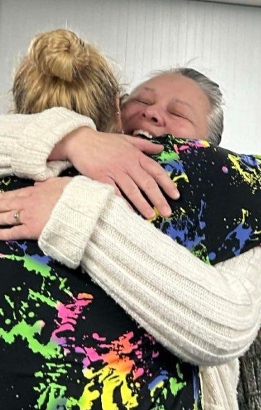 Two people hugging closely indoors, one in a black floral top and one in a white sweater.