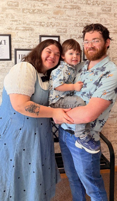 Three people smiling and hugging indoors against a brick wall.