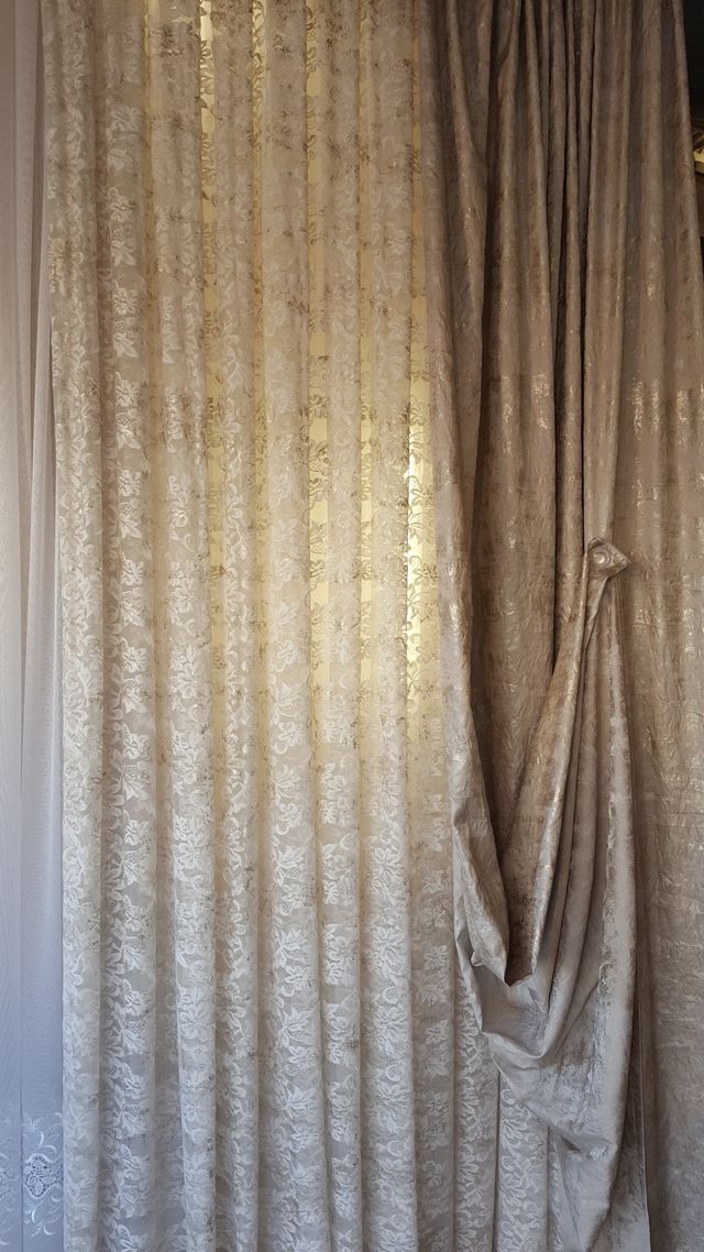 Tende plissettate beige con un fermatenda drappeggiato sulla destra, che coprono una finestra.