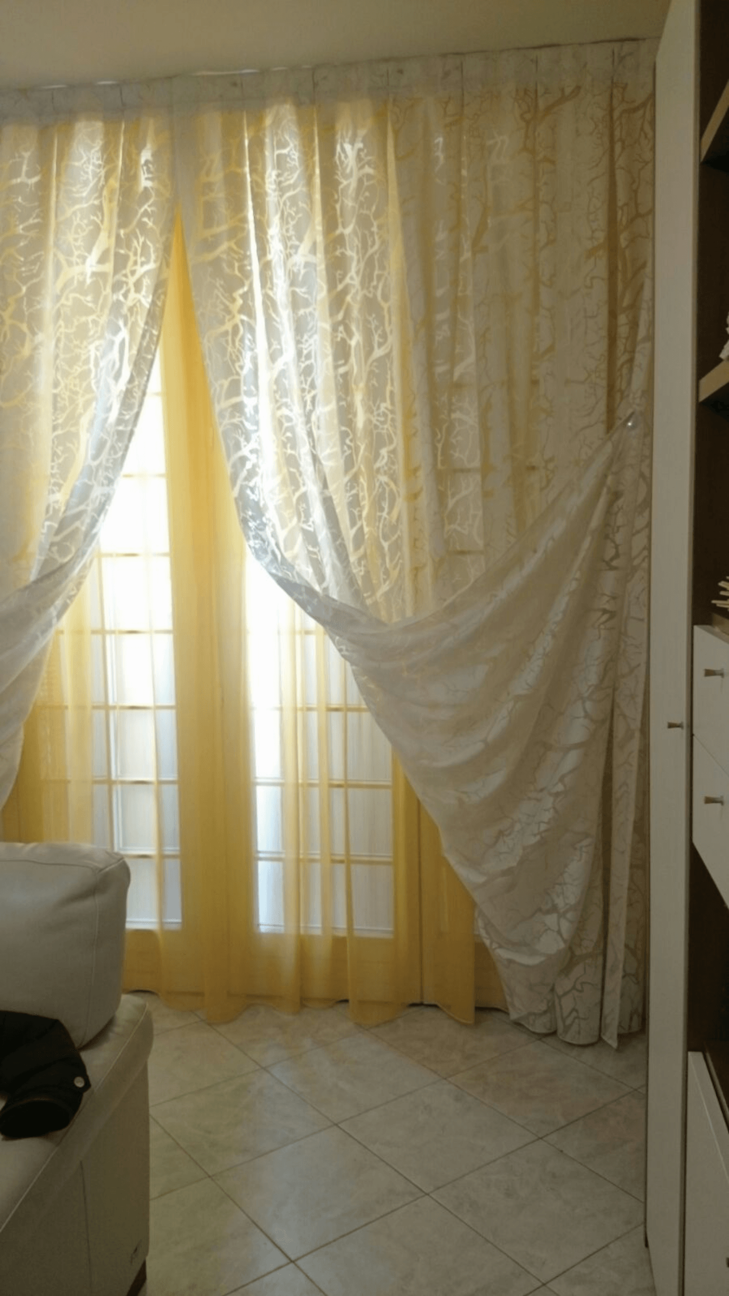 Tende trasparenti bianche e dorate su una finestra illuminata dal sole, con pavimento piastrellato e bordo del divano a vista.