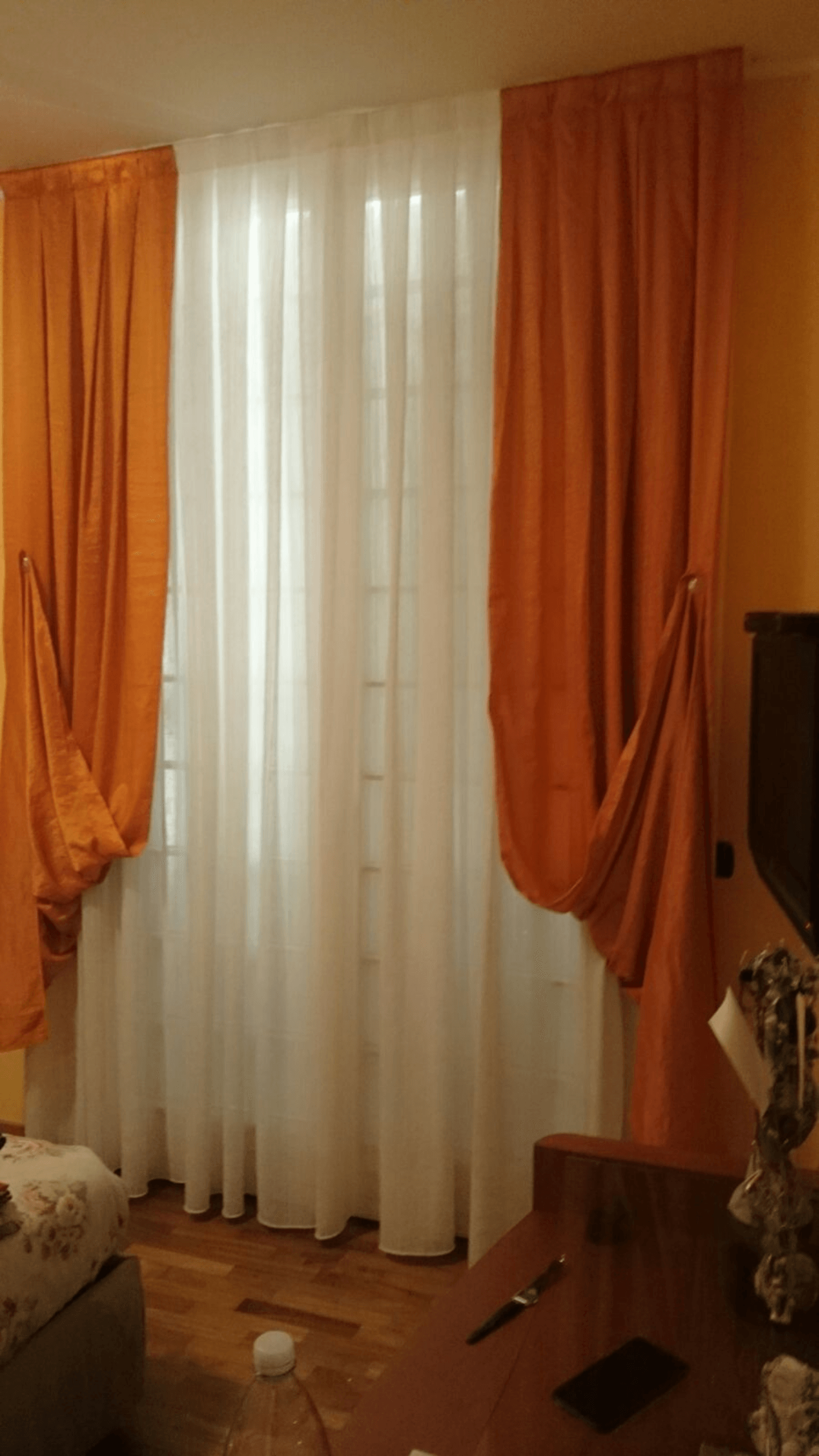 Soggiorno con tende arancioni che incorniciano veli bianchi accanto a un televisore e un tavolino in legno.