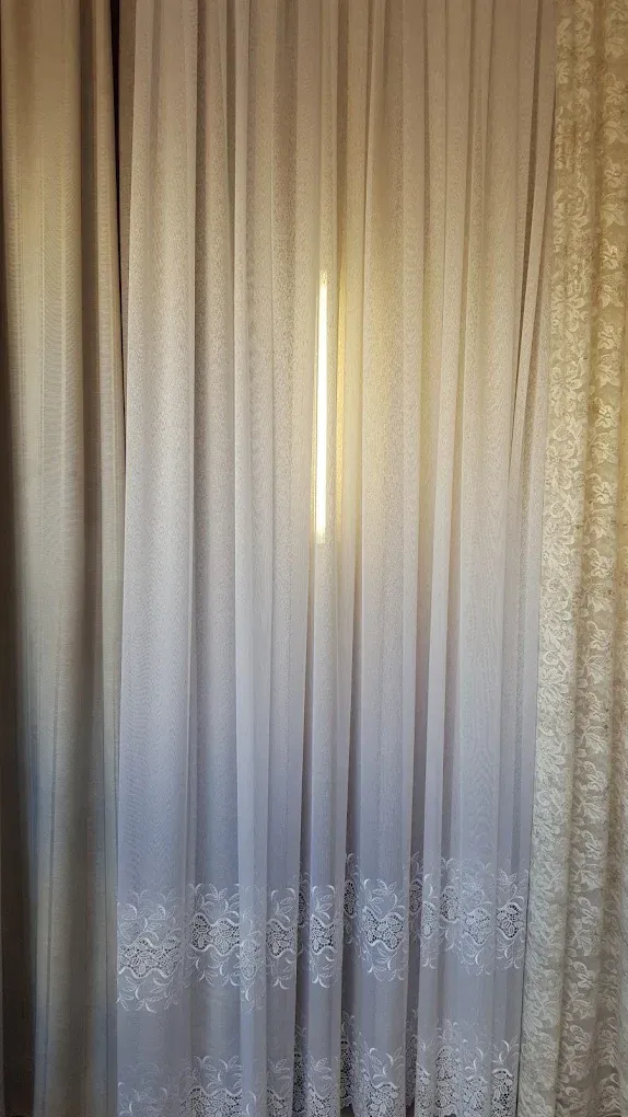 Tende beige trasparenti, con la luce del sole che filtra attraverso una stretta fessura al centro.