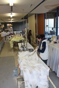 Laboratorio di sartoria con una persona che stira tessuti bianchi accanto a capi appesi e tavoli da cucito.
