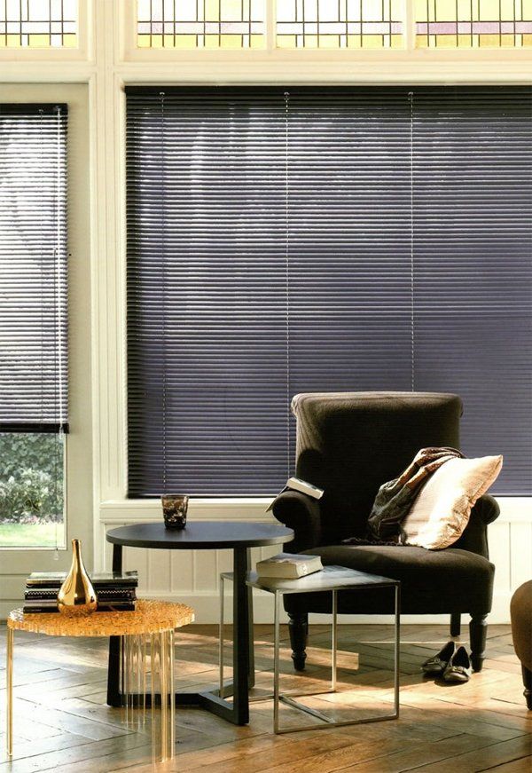 Soggiorno moderno con poltrona scura, tavolino, sgabello intrecciato e grandi finestre con tende.