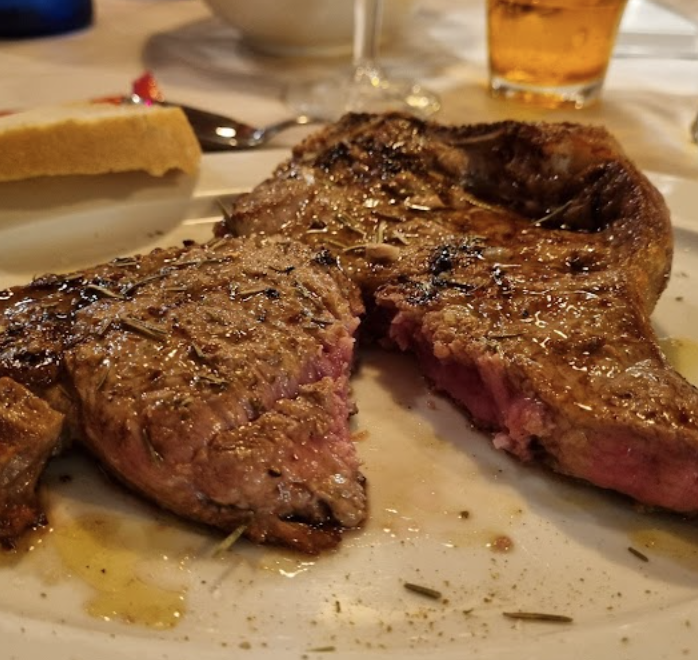 Una bistecca cotta, tagliata al centro e condita con erbe aromatiche, servita su un piatto bianco con un pezzo di pane accanto.