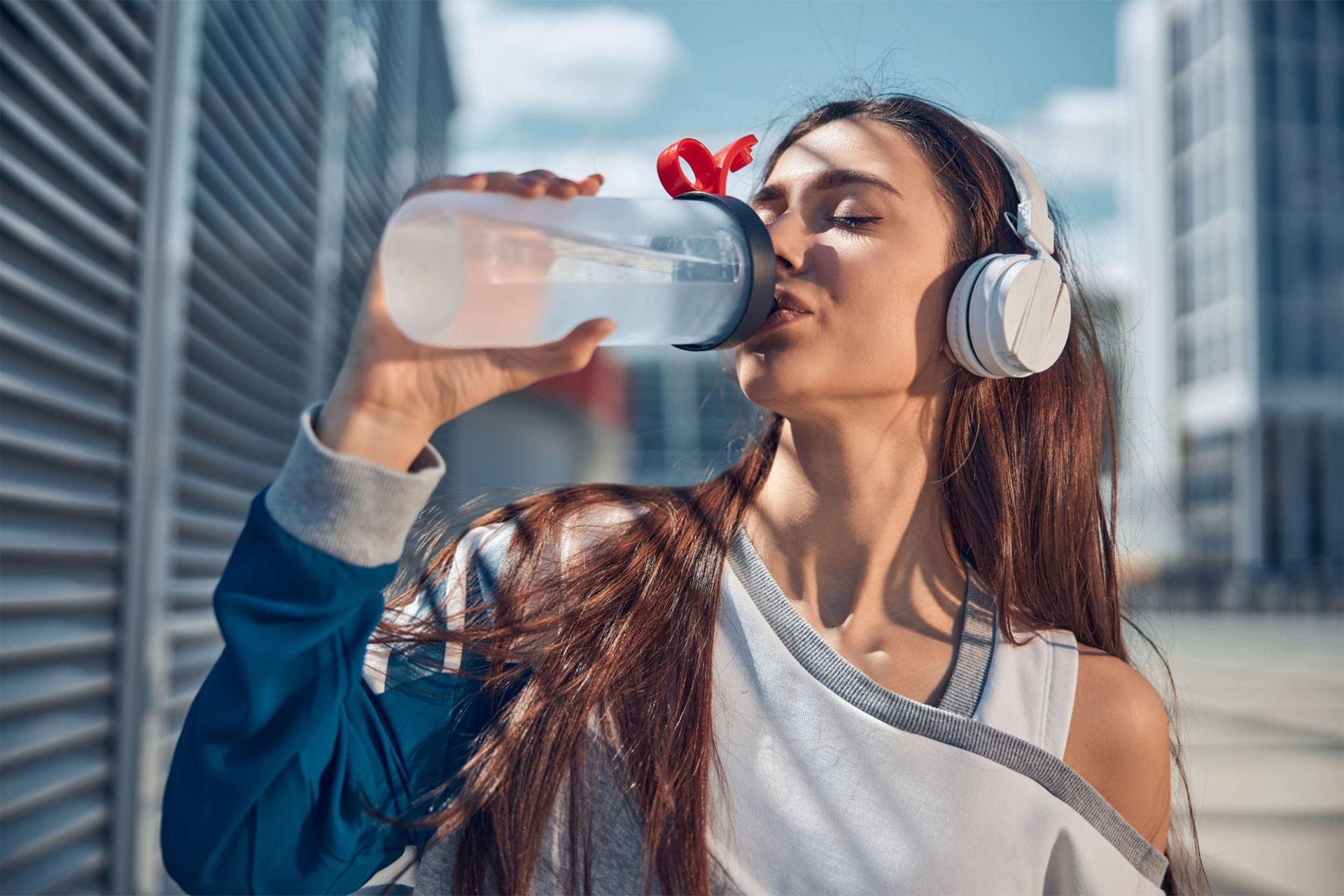 Person drinking water outdoors, wearing headphones, with city buildings in the background