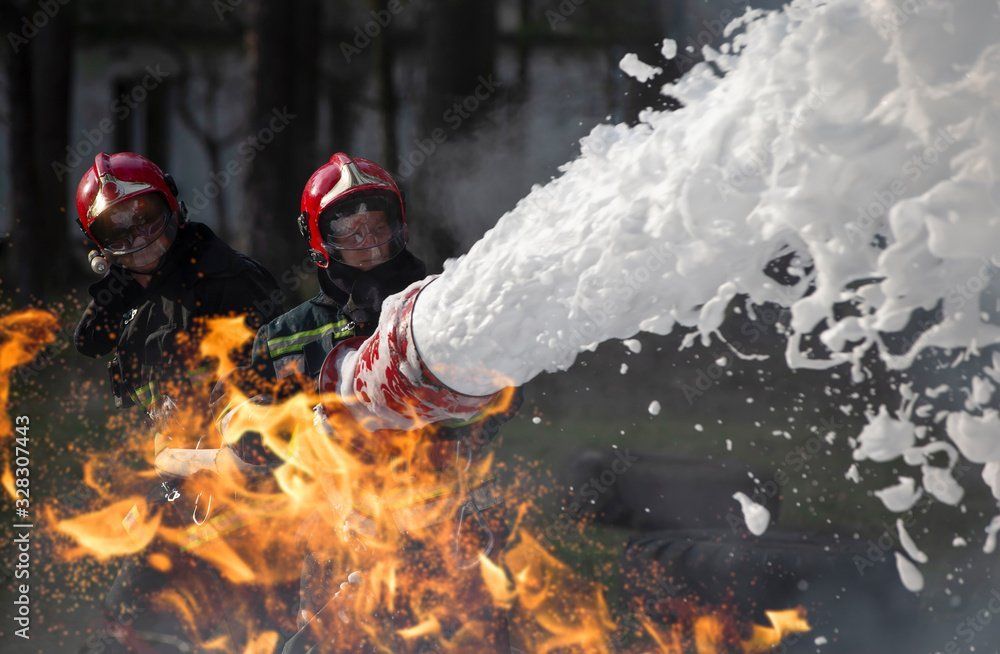 Firefighters using a foam hose to extinguish a fire; flames and foam are visible.