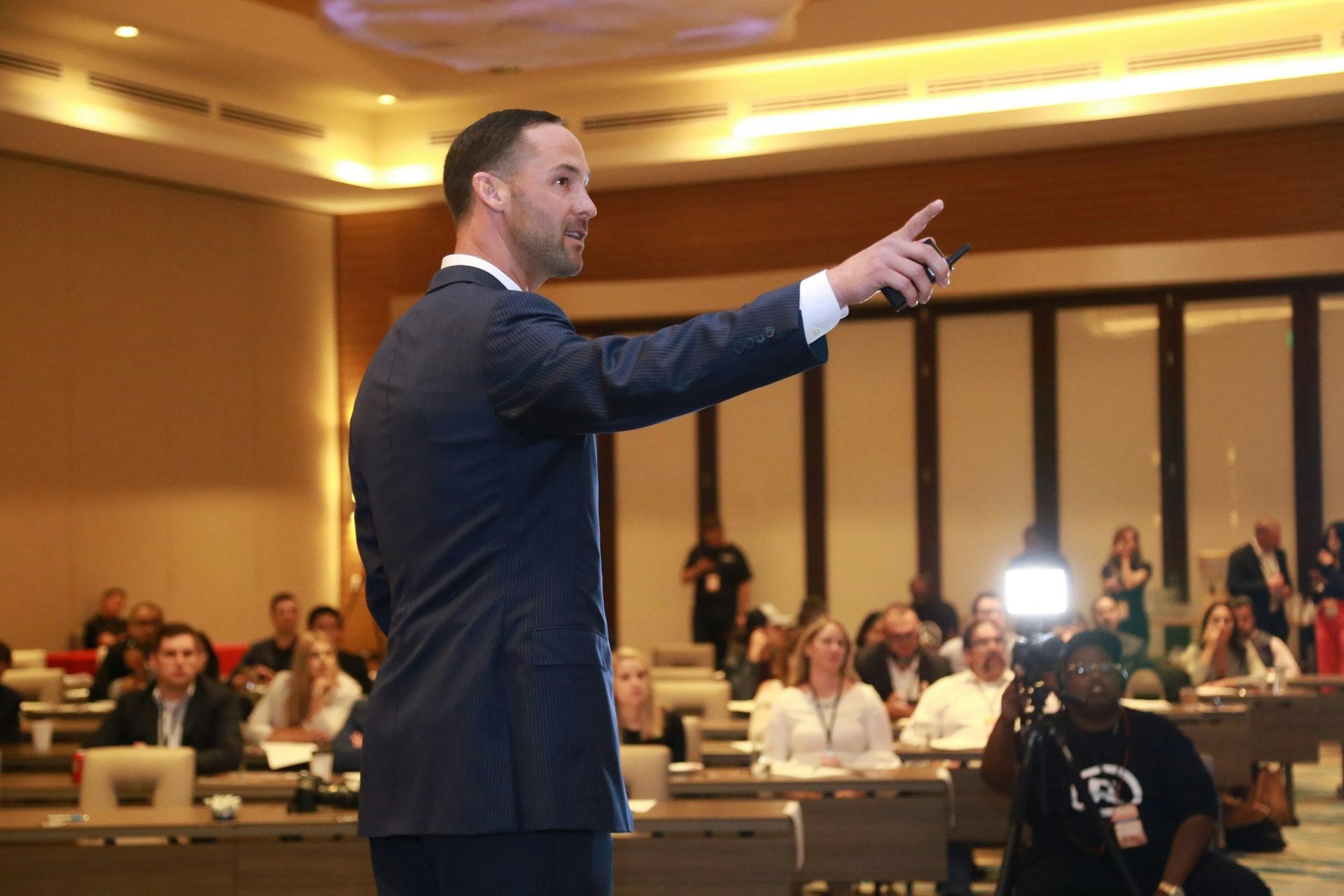 Man in suit giving a presentation to an audience in a conference room, gesturing with his hand.