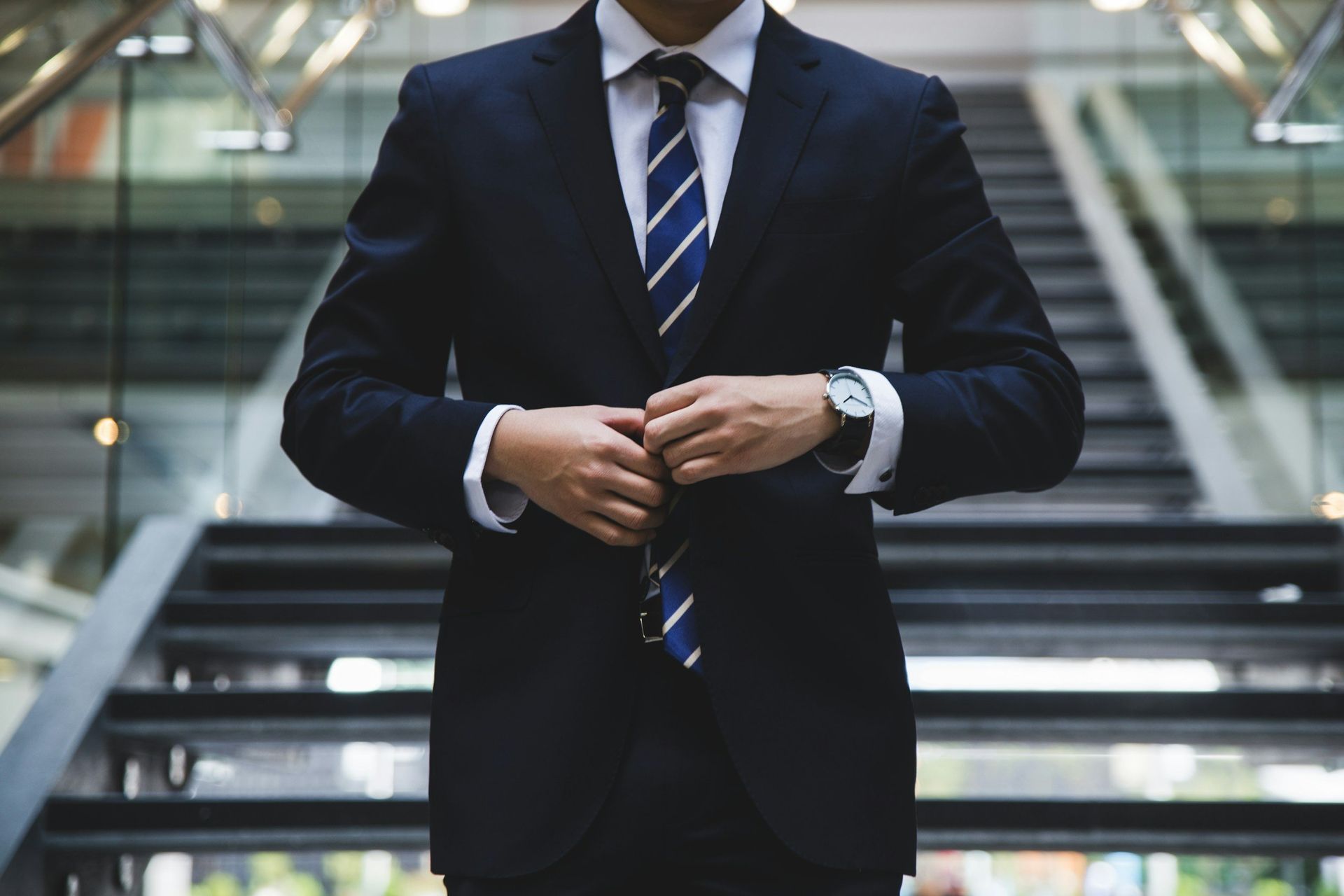 Man in a dark suit adjusting his jacket, standing in front of a staircase in a modern building.