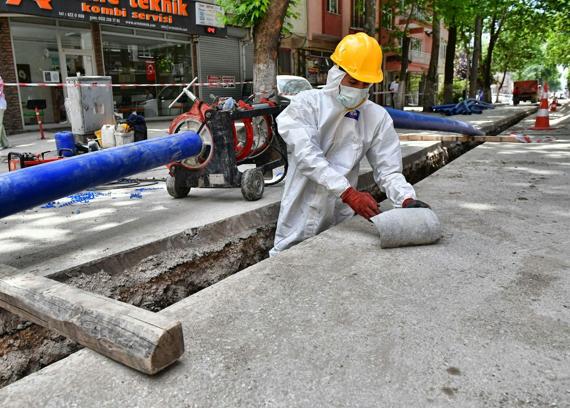 Construction worker in protective gear working in a street trench, next to pipes and equipment.